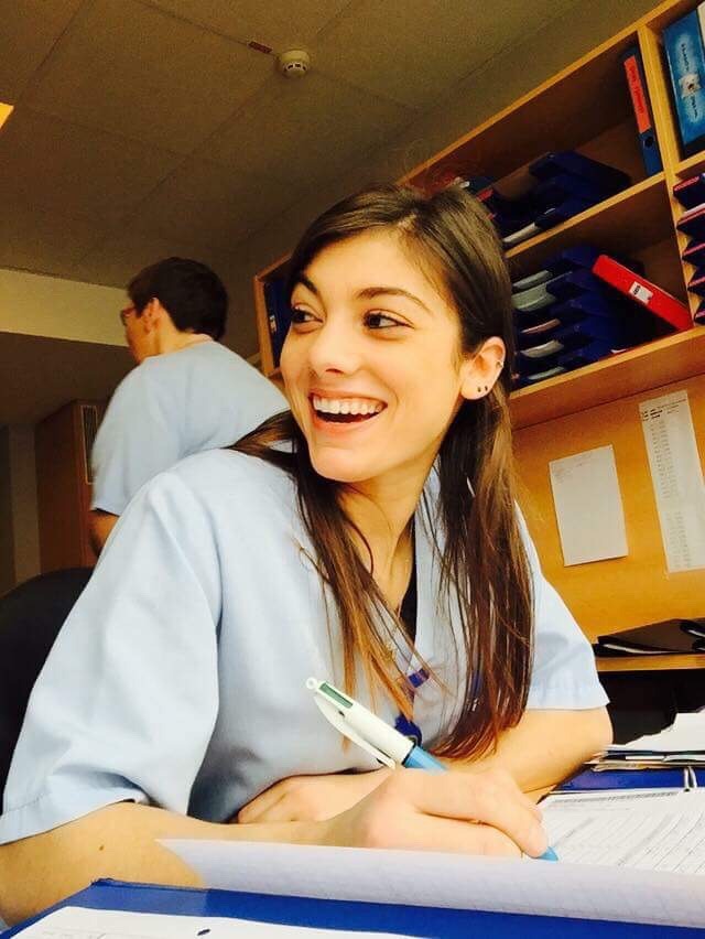 A smiling young female nurse in light blue medical scrubs writing notes at a hospital workstation.