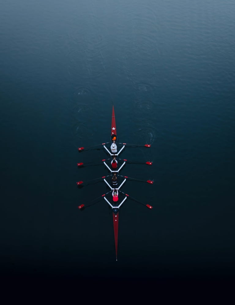 Aerial view of a four-person rowing crew in a red scull boat gliding on calm dark blue water.