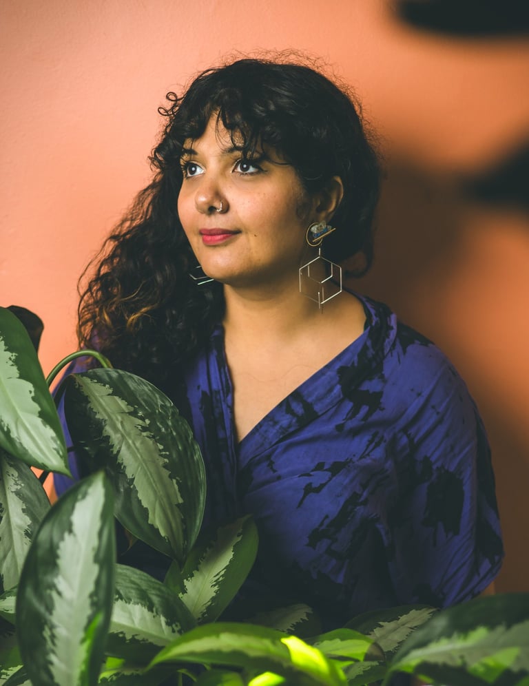 Portrait of Salima a brown skinned woman with long curly dark hair surrounded by plants. 