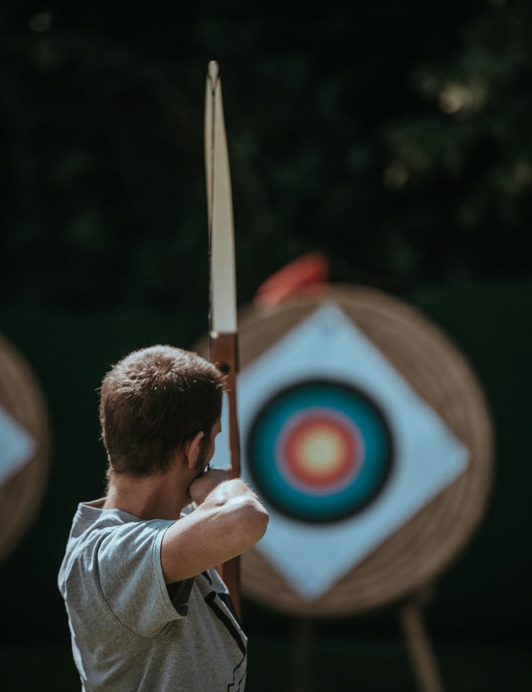 A male archer aiming a bow at a colorful target during outdoor archery practice.