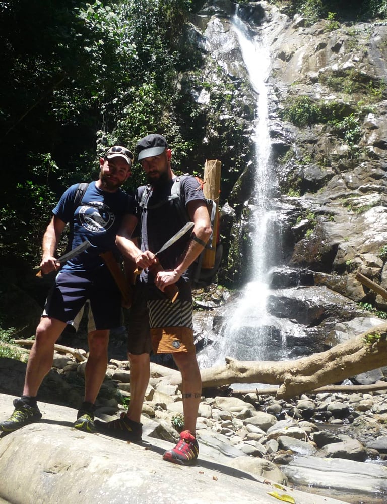 Two survival participants are standing in front of a waterfall holding a machete during the trip