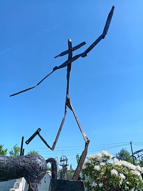 Tall abstract scrap metal sculpture of a human figure outdoors under a clear blue sky.