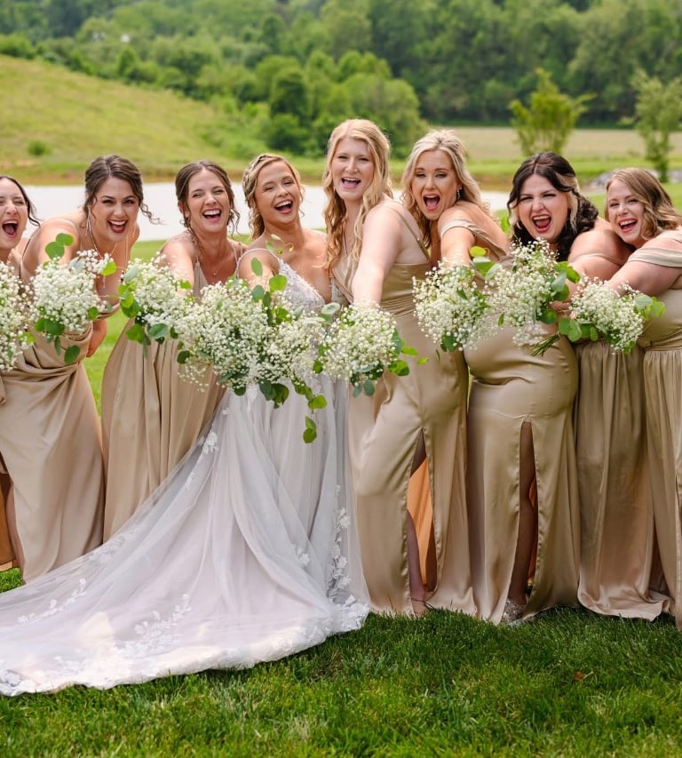 Bride surrounded by bridesmaids in gold dresses holding white bouquets, smiling outdoors, featuring wedding hair and makeup.