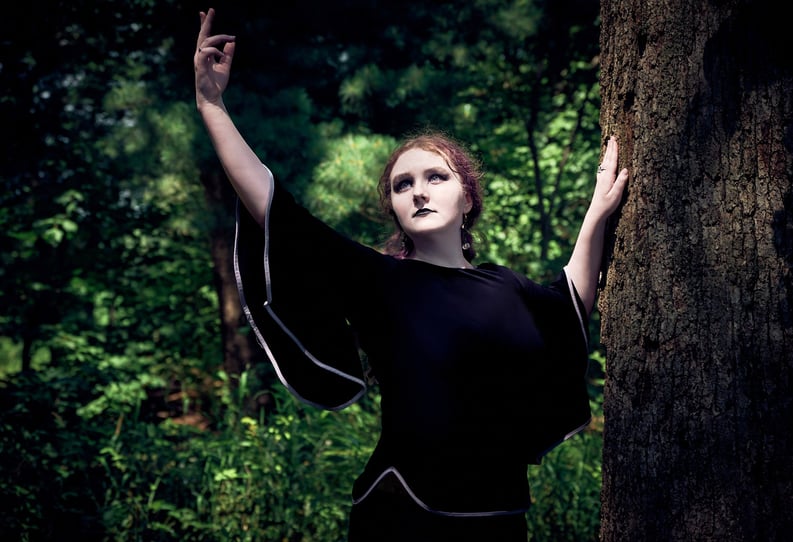 a woman in a black dress standing next to a tree dancing in the sunlight