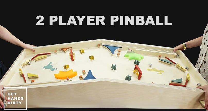 2 people standing in front of a 2-player pinball game table. 