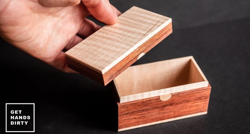 A hand holding a very small wooden box made out of maple, bubinga and leather.