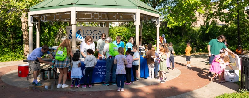 A bunch of children with a few adults approaching a table of adults in a park
