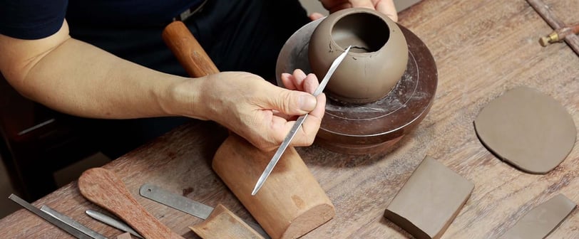 A hand holding a long-handled, pointed knife, carefully carving the edge of a teapot