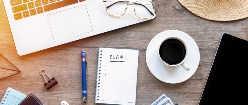 Uma caderneta de planos repousa numa mesa junto a notebook, lapiseira, xícara de café e óculos