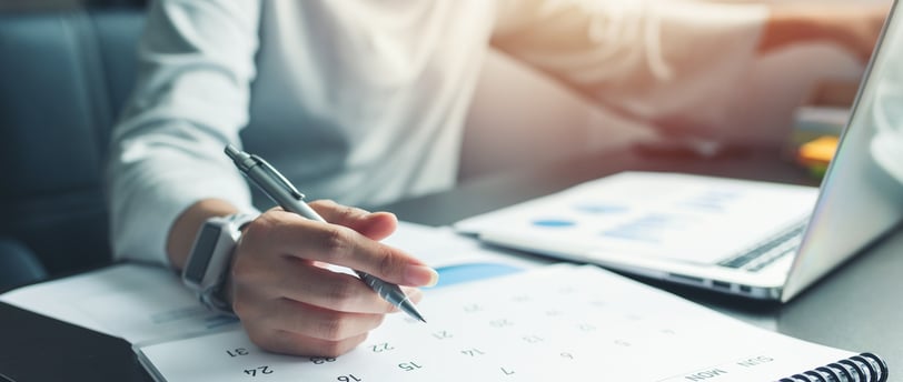 someone working at a desk with a calendar, laptop, and pen in hand