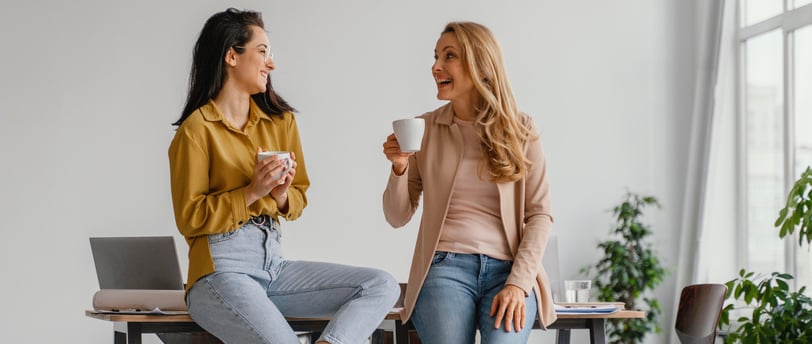 Two women talking and having coffee at a desk in an office, with a laptop on the table