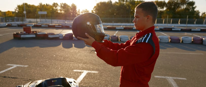 Jeune homme en combinaison rouge tenant un casque devant un karting sur une piste extérieure.