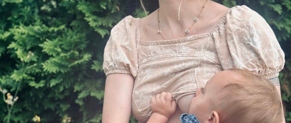 Mother sitting in front of a green bush, wearing a bow in her hair, holding and nursing her baby & looking away peacefully