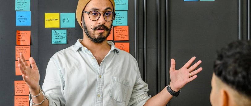 A casualy dressed man giving a talk standing infront of a board with colorful sticker notes on it