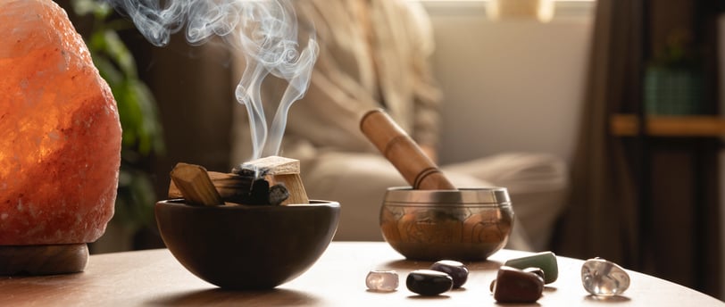 Table with crystals, palo santo, tibetan bowl, and a woman meditating in the background