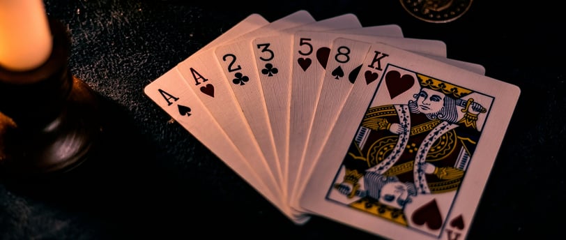 A spread hand of playing cards, witha pocketwatch, coin, chain and candlestick on the same surface in the background.