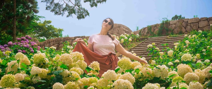 Une femme pose au milieu d’un champ d’hortensias blancs en fleur, sous un ciel bleu, sur les marches
