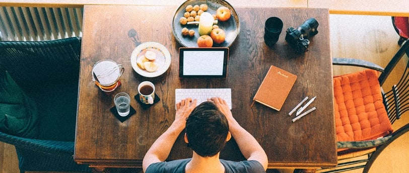 A top-down view of a writer working at a wooden table with a laptop, notebook, coffee, and snacks, surrounded by creative too