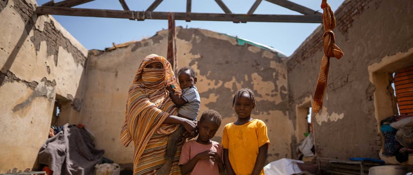 Sudanese woman and children standing inside a roofless home damaged by conflict.