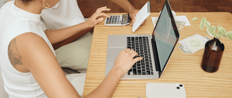 Couple working at a laptop with phone, papers, and calculator on a wooden desk