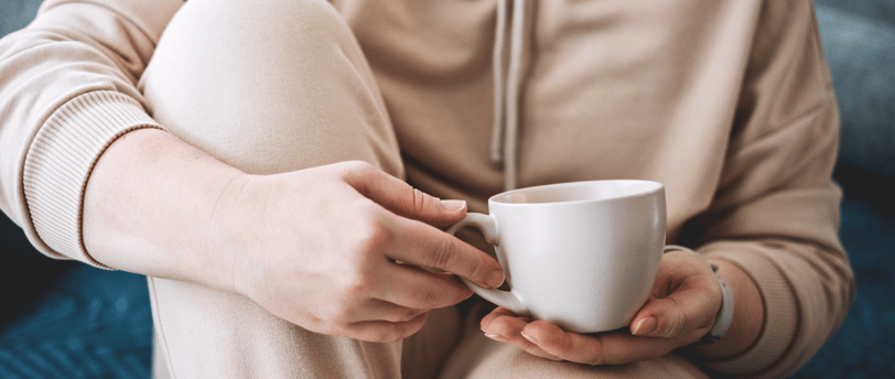 Person in loungewear holding a white mug while sitting with knees up on a sofa