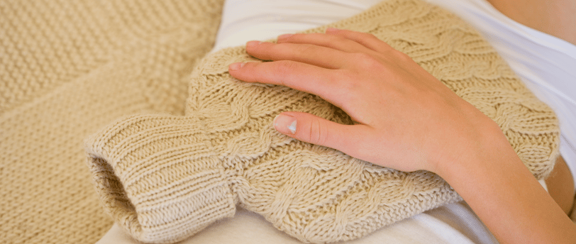 Person using a knitted hot water bottle on their lower abdomen, wearing cozy loungewear.