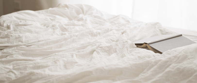 Unmade bed with soft white sheets and a closed journal on the mattress near a window.