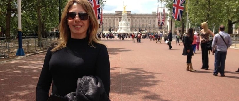 mulher loira, com óculos de sol, em frente a um palácio em rua com bandeiras britânicas