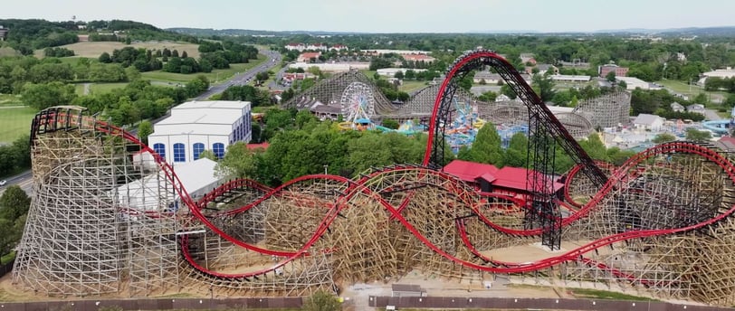 A black and red wooden roller coaster with several rides in the background