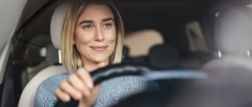 A person is seated in the driver's seat of a car, holding the steering wheel with one hand.