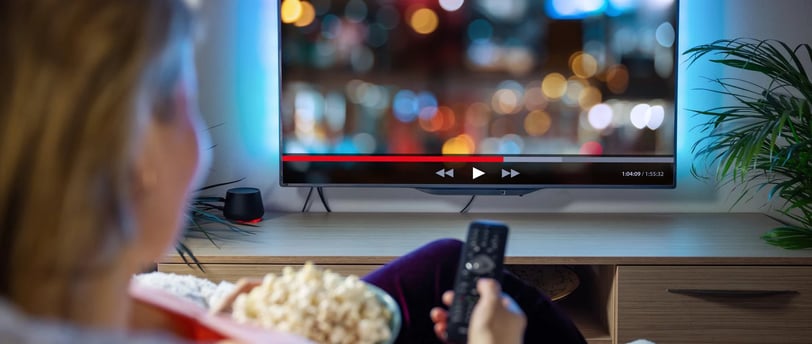 Uma pessoal segurando um balde de pipocas e com um controle remoto na mão assistindo a TV