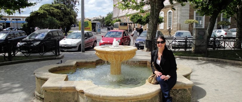 Foto da autora sentada numa fonte na praça próxima ao porto de Cádiz.