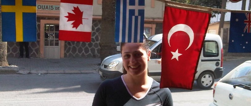 Foto da autora no centro de Izmir, entre uma bandeira turca e a movimentação local.