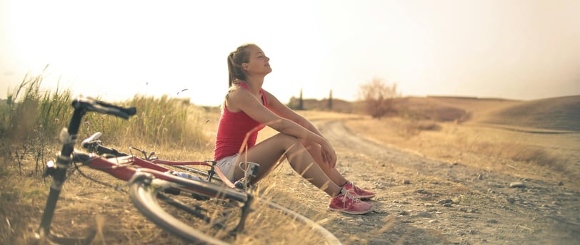 Woman with bicycle resting on countryside road in sunlight. Photo by Andrea Piacquadio on Pexels.
