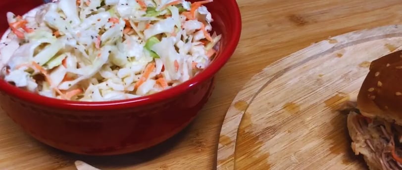 Creamy tangy and sweet coleslaw in a red bowl beside a pulled chicken sandwich on a plate.