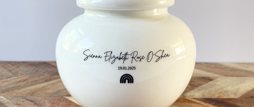 Rounded white ceramic urn with glossy finish, black script text “Sienna Elizabeth Rose O'Shea” and a small rainbow con.