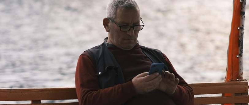 Homem sentado em um pier, camisa bordô e jaqueta verde, de óculos, olhando para o celular 