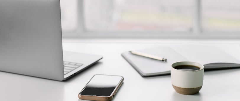a laptop, cell phone, notebook with pen, and a cup of coffee on a desk