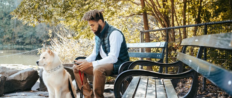 man sitting on park bench looking at phone holding the leash to a dog sitting beside him