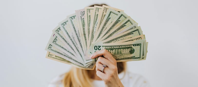 Person holding a fan of cash, displaying various U.S. dollar bills, covering their face.
