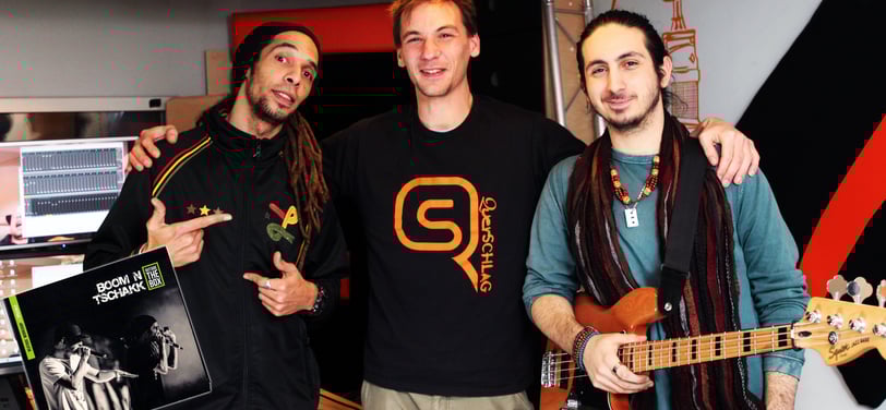 three men standing next to each other prducing songs, one is holding a guitar