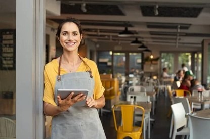 Uma empreendedora posando sorrindo na porta do seu restaurante, com um laptop nas mãos.