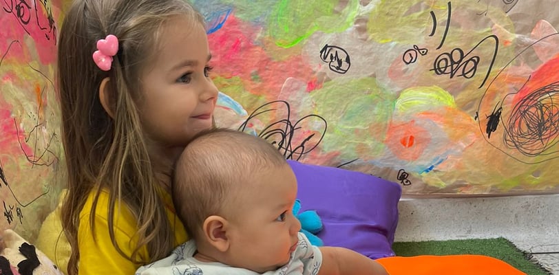 Niña abrazando bebe sentados entre cojines de colores vibrantes en Ukelele Jardín Infantil