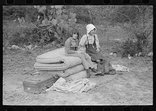 Two displaced girls sit on bundled bedding beside a suitcase during the Great Depression in Californ