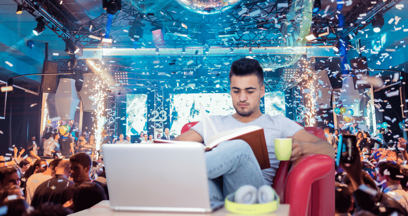 A man sitting in a red armchair holding a mug and reading a book with a party in the background