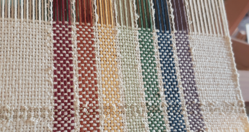 a loom with an unfinished hand-woven towel in muted rainbow pride flag colours