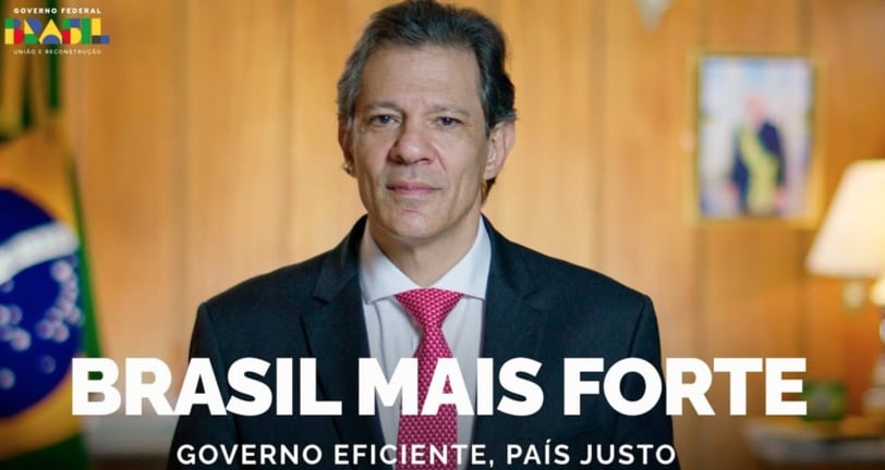Fernando Haddad, de terno e gravata grená, em gabinete, com a Bandeira e o retrato de Lula ao fundo