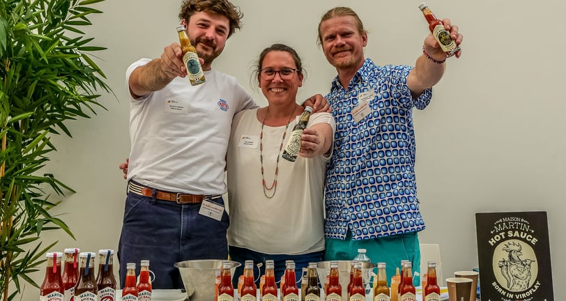 Deux hommes et une femmes qui tendent leurs bouteilles de sauces pimentées
