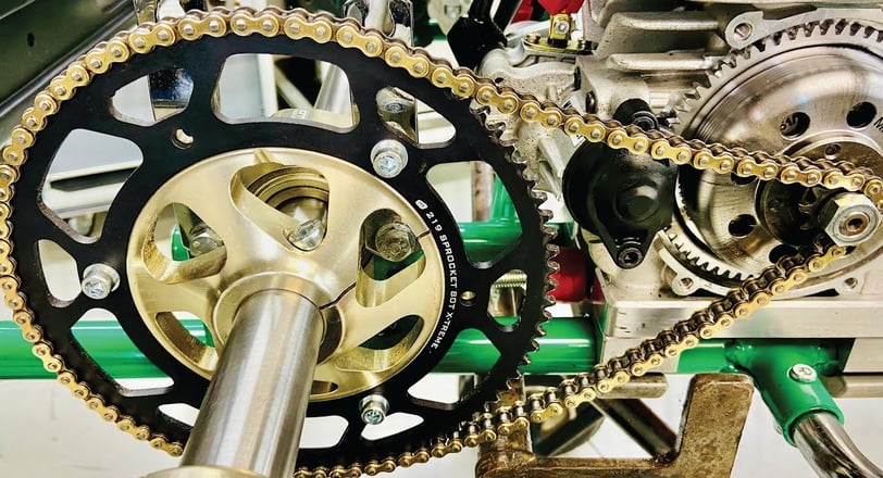 Chaîne dorée et engrenage robuste sur le système de transmission d’un kart, monté sur un axe métallique vert.