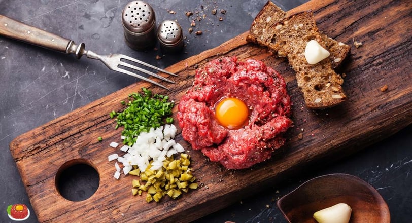 beef tartare on a wooden board, garnished with a raw egg yolk, diced onions, chives, pickles, rye bread, and a garlic clove